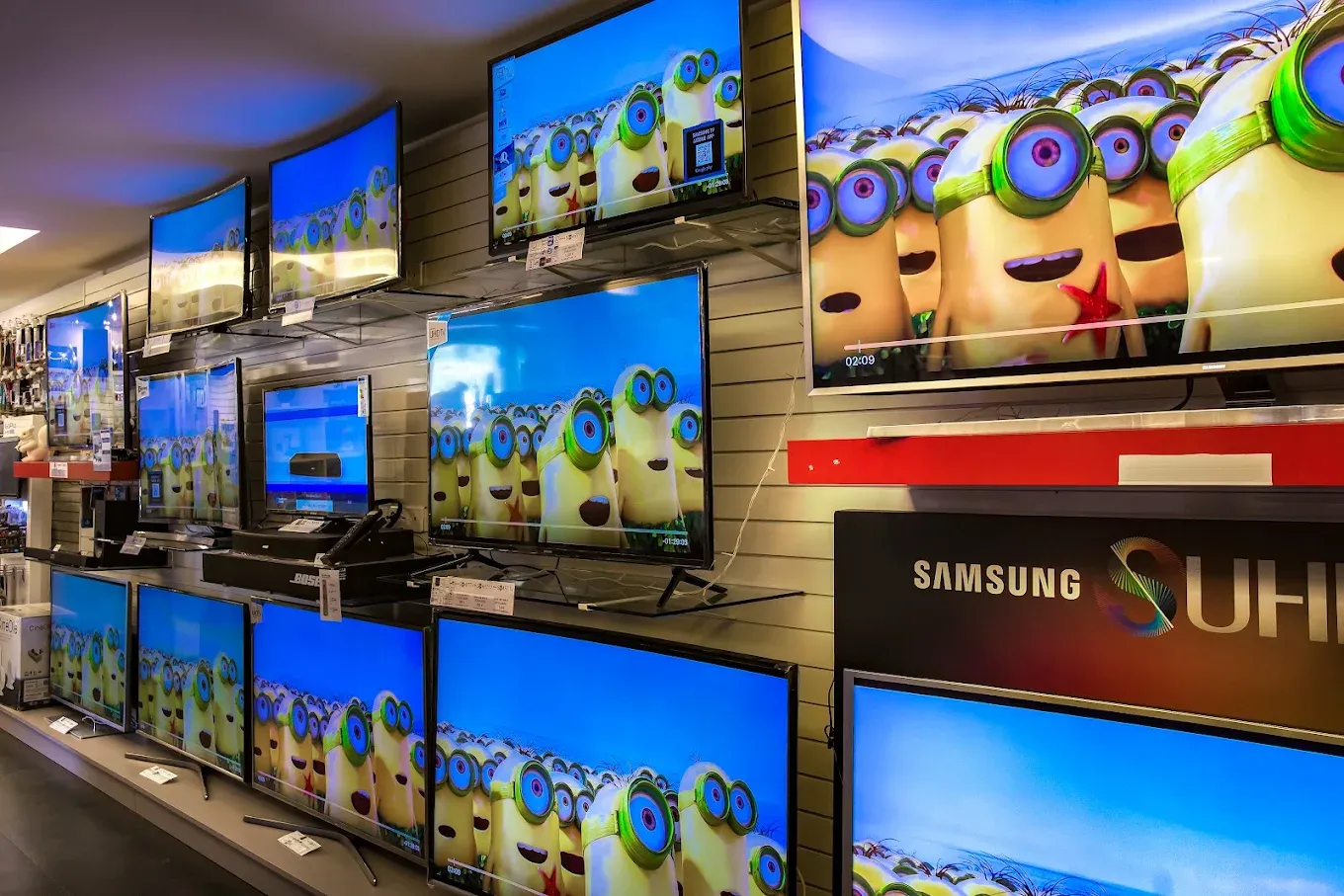 Mur de téléviseurs allumés, de plusieurs tailles, sous une enseigne Samsung, avec les barres de son en dessous.