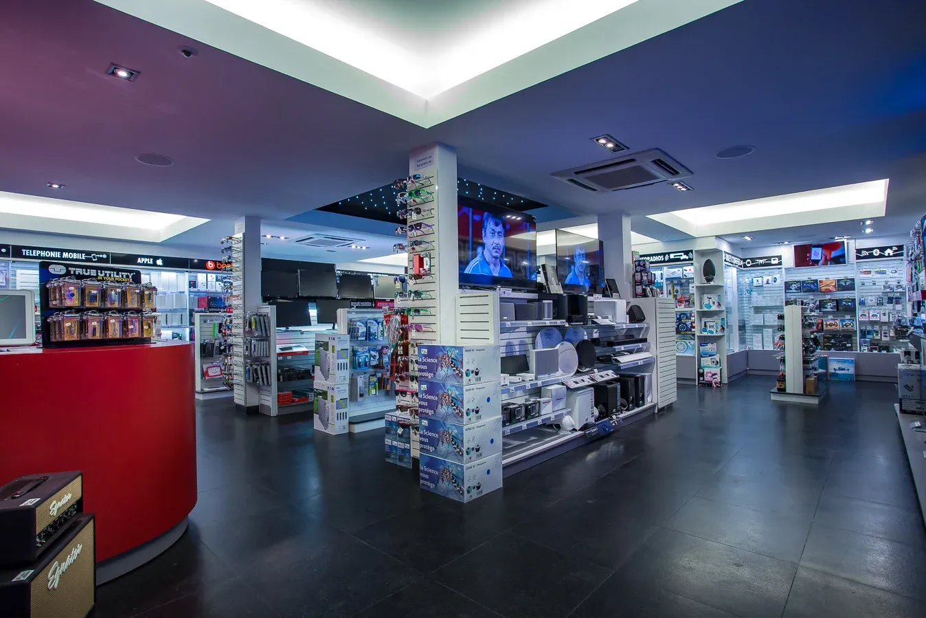 Vue d’ensemble du magasin : allées de présentoirs, comptoir rouge, murs d’accessoires et écrans allumés au fond.