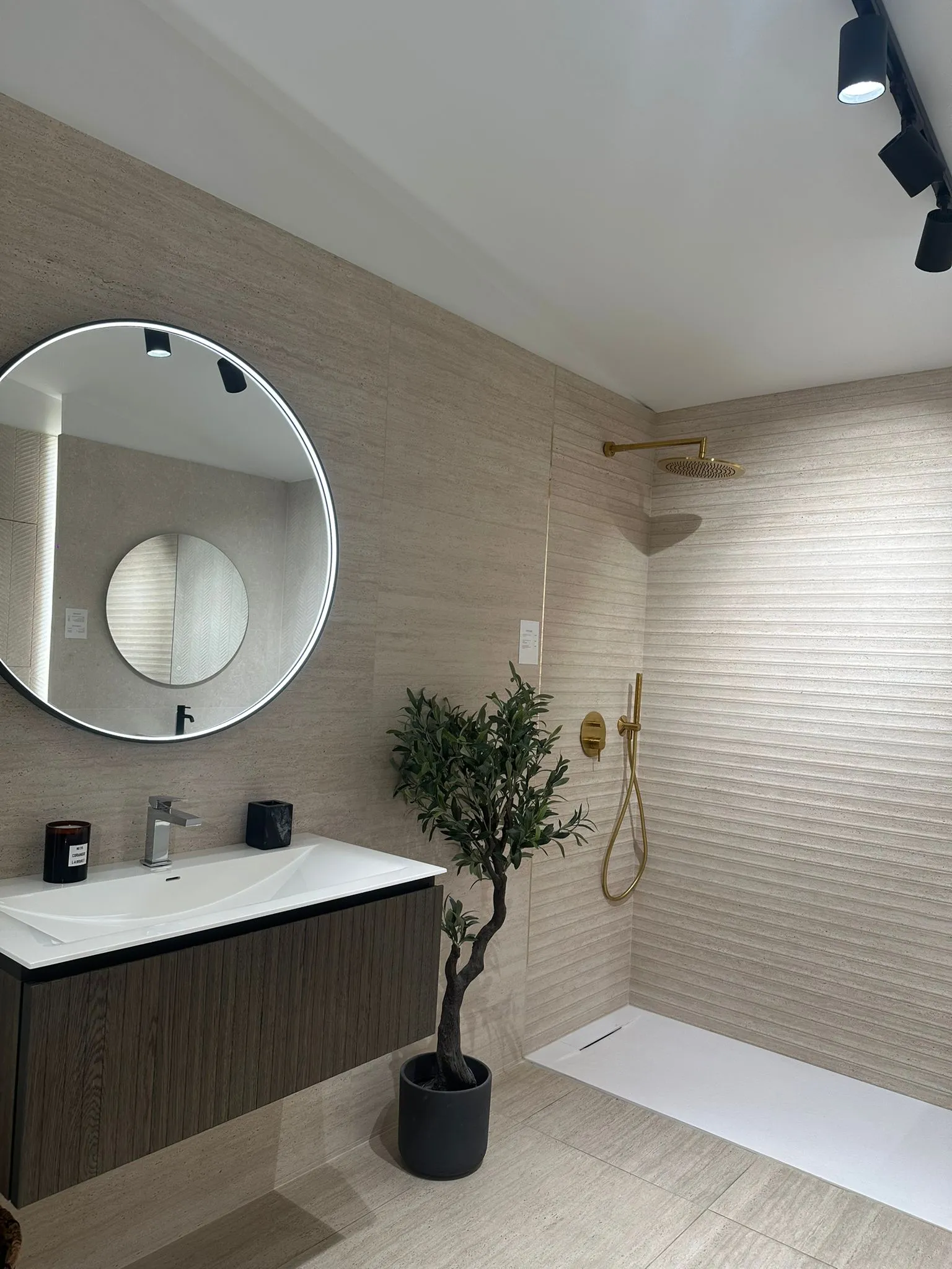 Bathroom in pale travertine: a large backlit round mirror, a wall-hung vanity unit, a potted olive tree and a walk-in shower with gold brassware.