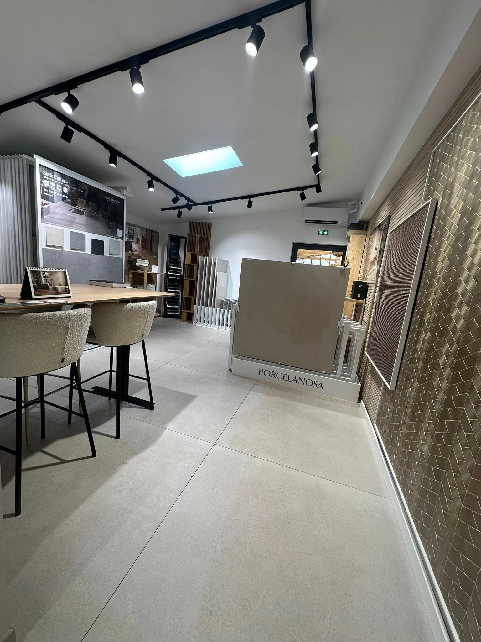 The showroom seen lengthways: high table and stools, a large-format ceramic slab on its display stand, gold mosaic wall to the right, skylight overhead.