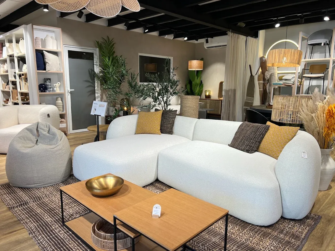 Espace salon en exposition : canapé d’angle en bouclé écru, table basse en chêne, pouf, tapis tissé et coussins ocre.